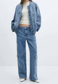 Lichtblauwe denimjas met elastische manchetten, gecombineerd met wijdvallende lichtblauwe jeans met cargozakken. Wit aansluitend topje eronder.