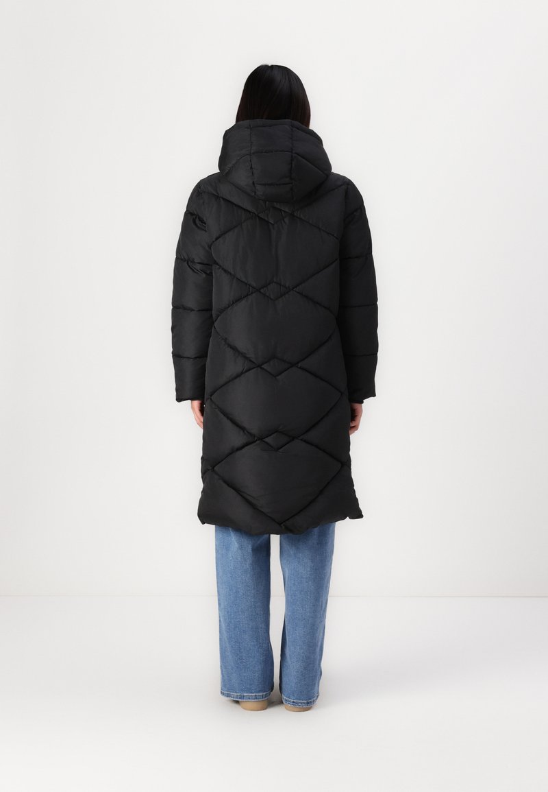 Manteau noir en duvet avec capuche, présentant un motif en losanges matelassé. Le manteau est de longueur genoux, associé à un jean ample bleu clair.