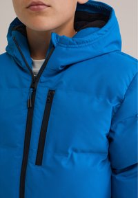 Chaqueta acolchada azul con un alto cuello y capucha, con cremalleras negras y un forro interior texturizado. Dos bolsillos con cremallera en la parte delantera.