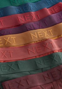Pile de ceintures de sous-vêtements pour hommes pliées en vert, bleu, rouge, violet, moutarde, orange et bordeaux, chacune embossée du texte "NEXT".