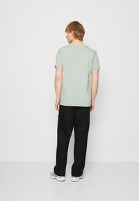 Šviesiai žalios medvilnės t-shirt marškiniai trumpomis rankovėmis, derinami su juodomis cargo kelnėmis. Modelis atsukęs nugarą, pasižymi minimalistiniu dizainu ir kasdiene išvaizda.