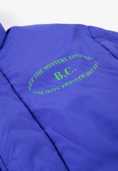 Tissu bleu avec un texte vert lisant "B.C." et "LET THE MYSTERY FIND YOU" dans une disposition circulaire sur un vêtement.