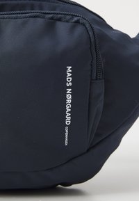 Mørkeblå bag laget av holdbart stoff, med glidelås. "MADS NØRGAARD COPENHAGEN" trykt i hvitt på siden.