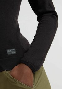 Maglia nera a maniche lunghe con texture a coste, caratterizzata da un'etichetta G-Star Originals cucita di lato, abbinata a pantaloni verde oliva.