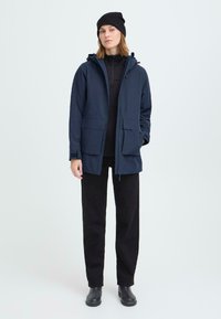 Jūras zila parka jaka ar kapuci, priekšējo rāvējslēdzēju un divām lielām kabatām. Uzvilkts virs melnā augsta kakla topa un melnām biksēm. Melna beanie cepure un zābaki.