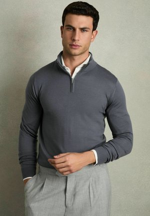 Suéter gris de manga larga con cremallera, de textura suave y diseño ajustado, combinado con pantalones plisados de un gris claro.