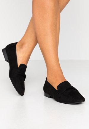 Ben iført sorte ruskind loafers med spidse tæer og lave hæle, stående på en lysegrå gulv mod en ensfarvet baggrund.