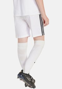 Weiße Sportshorts mit schwarzen Streifen, kombiniert mit knielangen weißen Socken und schwarzen Stollenschuhen mit einer durchsichtigen Sohle.
