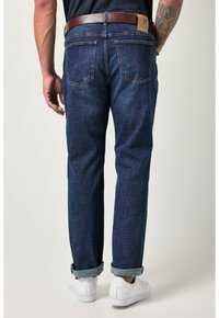 Jeans de mezclilla en azul oscuro con corte recto, puños enrollados y una etiqueta de cuero marrón en la parte trasera de la cintura. Zapatillas blancas visibles.