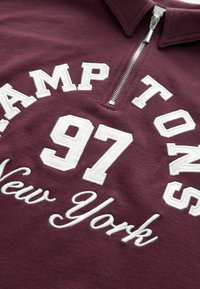 Sudor cu guler cu fermoar de culoare maronie, decorat cu text brodat alb: "HAMPTONS 97 New York." Material moale cu o textură fină.