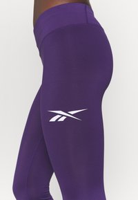 Lila leggings gjorda av stretchigt material med en slät yta. Vit Reebok-logotyp på sidan, hög midja med sömdetaljer.