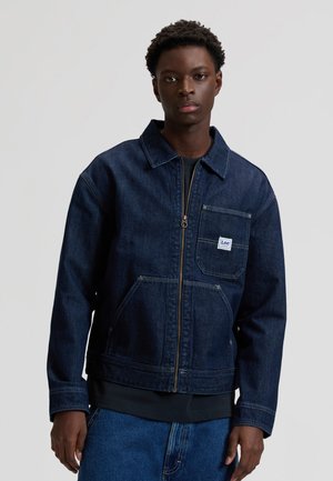 Ung mann iført en mørk blå denimjakke med glidelås, med en frontlomme og Lee-merke, kombinert med blå denimjeans.