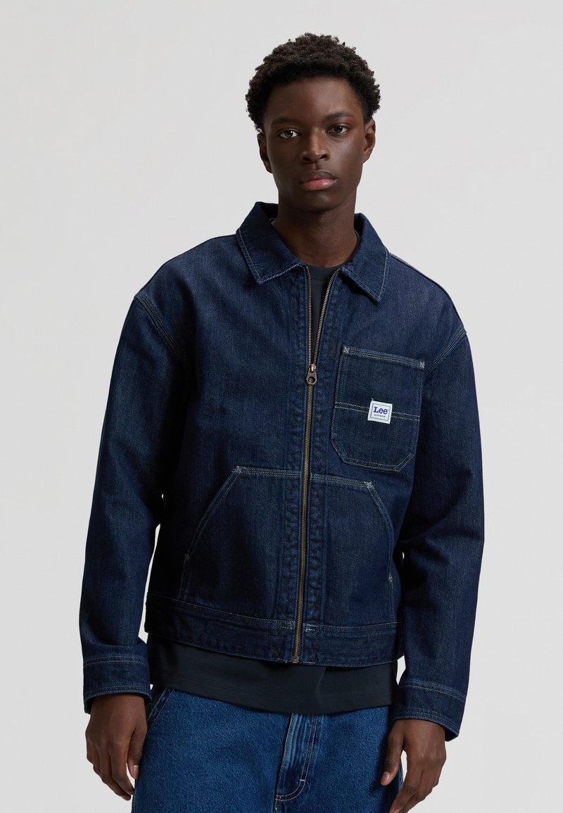 Νεαρός άνδρας που φοράει σακάκι από denim σε σκούρο μπλε με φερμουάρ και μπροστινή τσέπη, με ετικέτα της μάρκας Lee, σε συνδυασμό με μπλε τζιν παντελόνια.
