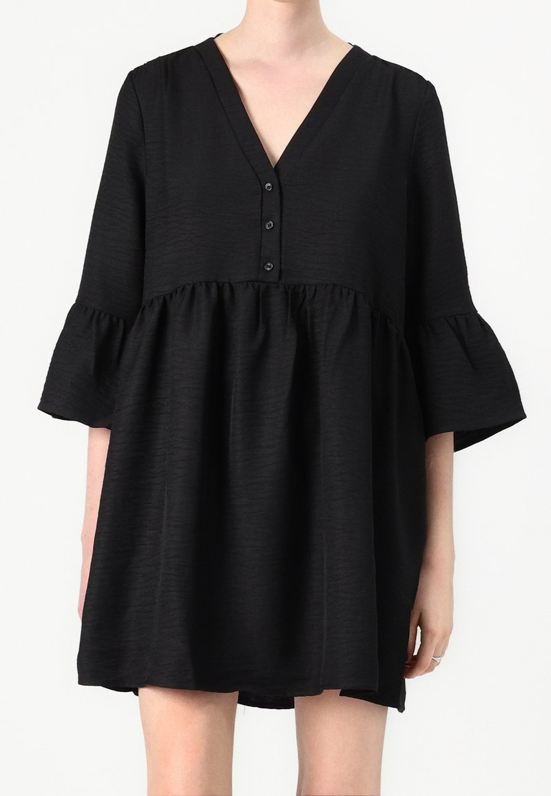 Femme portant une robe noire ample avec des manches trois-quarts à volants et un col en V avec trois boutons sur le devant.