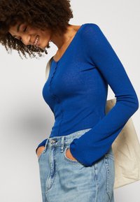 Blå, ribbestrikket langermet topp med rund hals og dekorativ knappdetalj, sammen med lyseblå denimjeans og en kremfarget skulderveske.