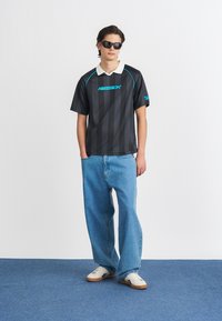 Melna svītrota īsām piedurknēm polo krekls ar baltu apkakli, kurā ir zils Reebok logo; apvienots ar platajām gaiši zilajām džinsām un baltām tenisa apaviem.