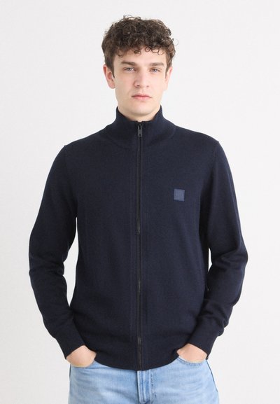 Sudadera de cremallera en color navy hecha de una tela suave, con cuello alto, mangas largas y un pequeño parche con logo en el lado izquierdo del pecho.