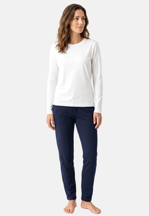 Frau steht barfuß, trägt ein weißes Langarmshirt und marineblaue Hosen und schaut leicht zur Seite auf einem einfarbigen Hintergrund.