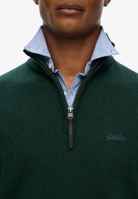 Mørkegrøn strikket zip-sweater med en struktureret overflade, med en lagdelt lyseblå stribet skjorte under og mærke-zip.