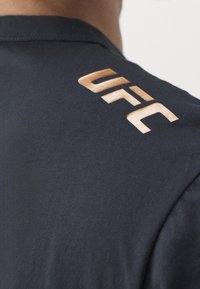 Ramiona i górna część pleców osoby w czarnej koszulce z miedziano kolorowym logo "UFC" w pobliżu dekoltu.