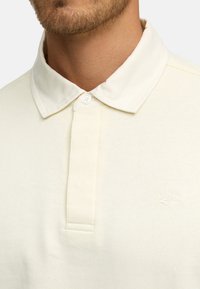 Lyse beige polo-skjorte med klassisk krage, treknaps halslinje og subtil teksturert stoff med preget logo.
