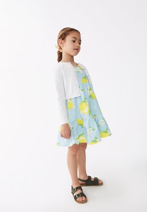 Vestido azul claro con estampado de limones y flores, falda de capas, combinado con un cárdigan blanco corto y sandalias oscuras.