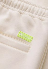 Pantalons de survêtement crème avec un tissu doux et texturé, une taille froncée et un patch logo 'LEVEL UP' vert fluo sur la poche.