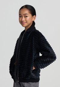 Giacca bomber in velluto nero con texture a coste, chiusura con zip e tasche laterali. Presenta un colletto e polsini a coste neri a contrasto.