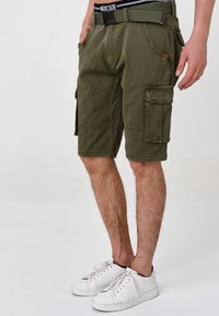 Shorts cargo vert olive avec plusieurs poches, une ceinture et une finition texturée. Associés à des baskets blanches sur un fond uni.