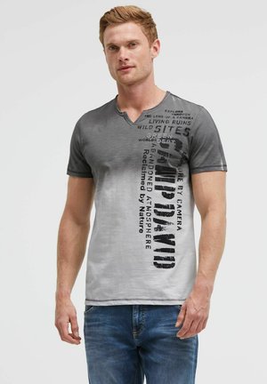 Man wearing gray and white gradient short-sleeve shirt with black vertical text and blue jeans, standing against a plain background.