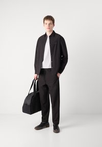 Chemise noire à boutons sur un t-shirt blanc, pantalon noir et baskets noires. Tenue avec un sac de sport noir. Design simple et moderne.