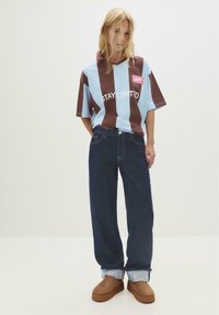 En blond person iført en blå og brun stribet skjorte med teksten "STAY UNITED", mørke oprullede jeans og brune slip-on sko, står foran en hvid baggrund.