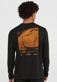 Must musta särk mustade pikkade varrukatega, mille seljal on oranž graafika, mis kujutab rannamaastikku ja teksti "O'Neill Surf Shop" asukohtade kohta.