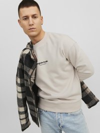 Lichtbeige sweatshirt met ronde hals en zwart logo. Grijs en zwart geruit jack over de schouder gedrapeerd. Blauwe denim jeans.