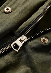 Tessuto verde oliva con una superficie liscia, dotato di una cerniera metallica e un bottone a pressione. Nastro della cerniera nero e dettagli in metallo argentato visibili.