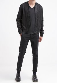 Svart bomberjacka med en jämn textur, kombinerad med en åtsittande svart t-shirt och smala svarta byxor. Svarta skor fulländar outfiten.