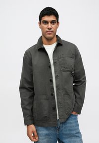 Σκούρο γκρι σακάκι με κουμπιά, γιακά από βελουτέ ύφασμα, τσέπη στο στήθος και δύο μπροστινές τσέπες, κατασκευασμένο από ανθεκτικό ύφασμα, φορεμένο πάνω από ένα ανοιχτόχρωμο πουκάμισο.