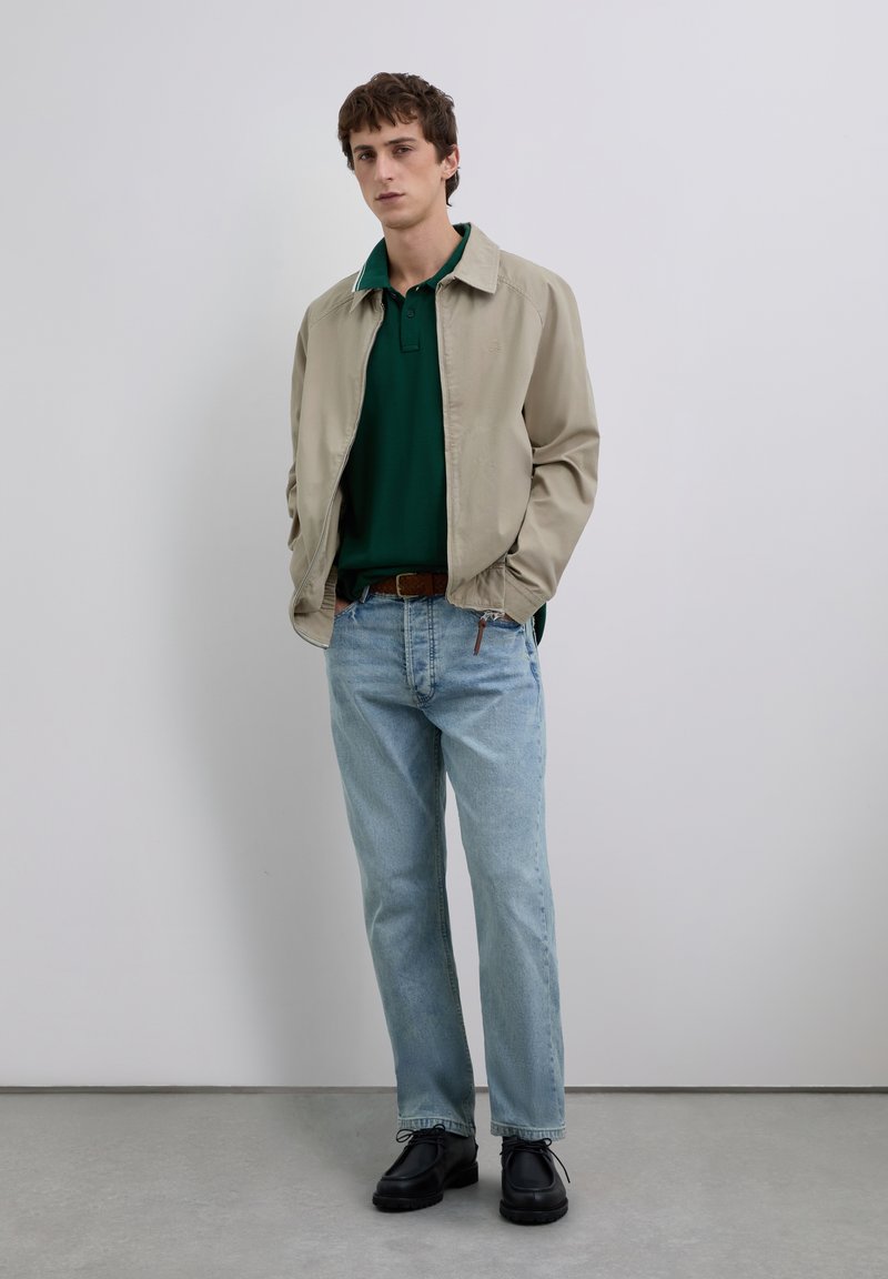 Joven vestido con una chaqueta beige, polo verde oscuro, jeans azules claros, cinturón marrón y zapatos negros con cordones, de pie contra un fondo liso.