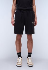 Black sweatshorts with a drawstring waist, side pockets, and a small logo on the left leg. Paired with light beige and white sneakers.