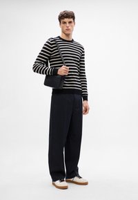 Jersey a rayas en blanco y negro con puños acanalados, pantalones negros y zapatillas ligeras con suelas marrones. Modelo sosteniendo un bolso cruzado negro.