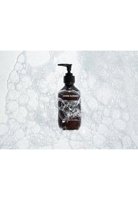 Flacon pompe sombre étiqueté "Grown Alchemist" entouré de bulles de savon blanches sur un fond clair.