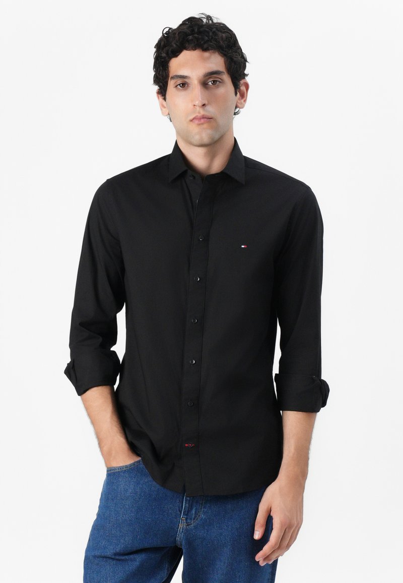Black button-up shirt with a collar, long sleeves rolled at the cuffs, and a small logo detail on the chest. Smooth, woven fabric.