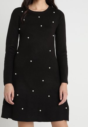 Schwarzes, langärmliges Strickkleid mit verstreuten kleinen weißen Perlenverzierungen, getragen von einer stehenden Person vor einem einfarbigen Hintergrund.