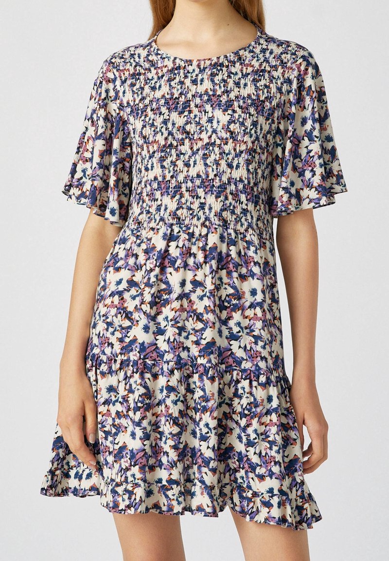Robe courte fleurie avec un corsage smocké, des manches à volants et une jupe à étages. Les couleurs comprennent des motifs floraux crème, violet, bleu et rose.
