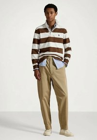 Młody mężczyzna stoi w brązowej i białej pasiastej koszulce rugby na niebieskiej koszuli zapinanej na guziki, beżowych spodniach i jasnobeżowych trampkach.