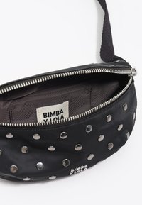 Czarna materiałowa torba na ramię z srebrnymi ćwiekami, zamknięciem na zamek i wewnętrzną metką. Charakteryzuje się gładką fakturą i zaokrąglonym kształtem, zaprojektowana z myślą o uniwersalności.