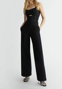 Mono negro con un corsé ajustado que presenta un corte, detalle con cordones en la cintura y pantalones de pierna ancha. Combinado con sandalias de tacón brillantes.