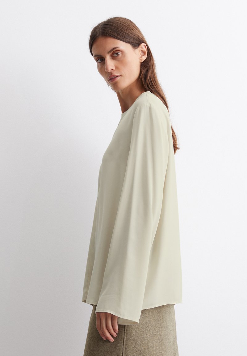 Hellgrüne Bluse mit langen Ärmeln, rundem Halsausschnitt und glatter Textur. Getragen mit einem beigen Rock, minimalistisches Design, ohne Muster.