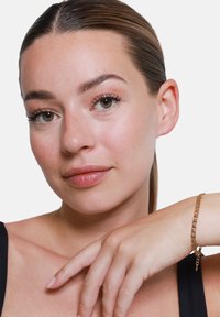 Bracciale in oro con maglie intrecciate, appoggiato sulla pelle con una tonalità neutra e liscia. La mano si appoggia delicatamente sul polso.