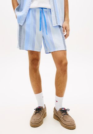 Hombre con pantalones cortos de rayas azules y blancas con cordón azul, calcetas blancas y zapatos de ante marrón sobre un fondo liso.